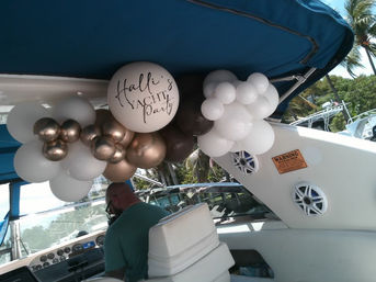 Cluster of white, metallic bronze, and brown balloons under a blue canopy on a yacht, featuring a large white balloon scripted Yacht Party, hanging above the helm and white seating with speakers and palm trees visible at the marina.