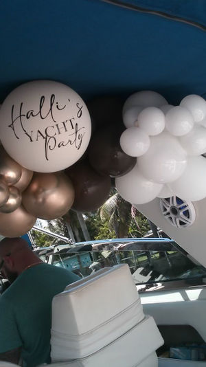 Metallic gold, white and dark balloons forming a garland with a personalized print above a motor yacht helm under a blue bimini, palm trees and boats at a sunny marina.