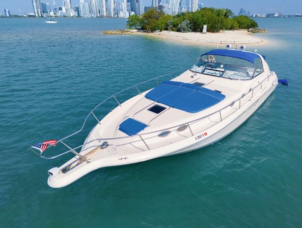 Luxury white motor yacht with blue sun pads and bimini, American flag at the bow, anchored in turquoise water near a small sandy island with greenery and a distant city skyline — sunny coastal scene.