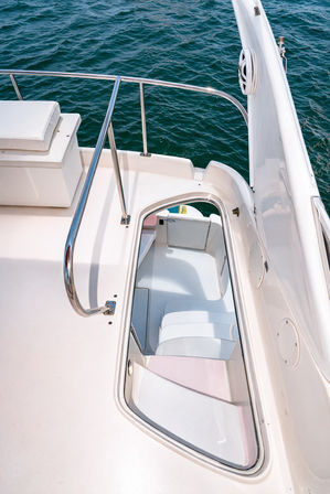 White yacht deck with polished stainless railing overlooking deep teal open water, hatch open to a sunlit cockpit with white cushioned seating — boat, deck, and ocean scene.