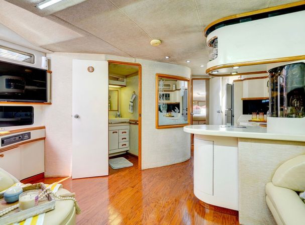Bright luxury yacht salon with curved white galley island, glossy wood floors, built-in TV, cream leather seating, and an open door to a compact bathroom