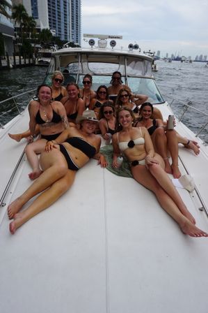 Group of women in swimsuits lounging and smiling on the bow of a yacht during a waterfront city boat party, skyline and water in the background.