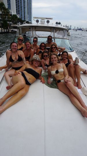 Group of women in bikinis relaxing on the bow of a white motor yacht near a city marina and skyline, smiling and holding drinks on a cloudy day.