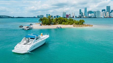 White motor yacht anchored near a small palm-covered tropical sandbar in turquoise water, with jet skis and a distant coastal city skyline and bridge under a partly cloudy sky