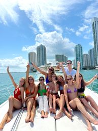 Eight friends in colorful swimsuits lounging and cheering on the bow of a white yacht, holding drinks with turquoise water and a high-rise waterfront skyline under bright blue skies.