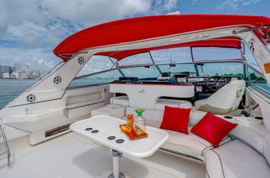 Luxury white yacht deck with red canopy, white leather seating and red throw pillows around a table with glass decanters, cruising past a coastal city skyline under a partly cloudy sky.