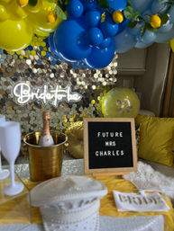 Cheerful indoor bridal shower setup with blue and yellow balloon garland, neon bride to be sign on a gold sequin backdrop, champagne in a gold ice bucket, and a letter board reading Future Mrs on a decorated table