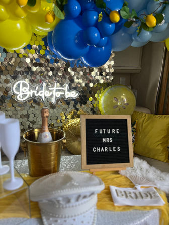 Cheerful indoor bridal shower setup with blue and yellow balloon garland, neon bride to be sign on a gold sequin backdrop, champagne in a gold ice bucket, and a letter board reading Future Mrs on a decorated table