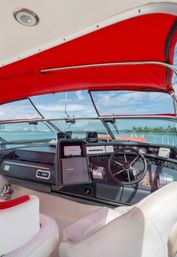 Yacht helm under a bright red bimini canopy - leather captain's seats, steering wheel and instrument panel with navigation screens, looking out over turquoise water and a distant coastal skyline on a sunny day.