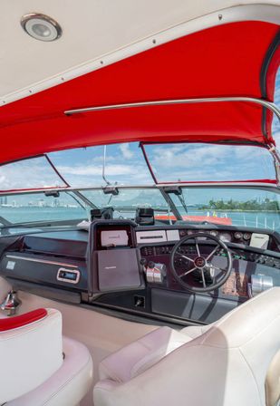 Yacht helm under a bright red bimini canopy - leather captain's seats, steering wheel and instrument panel with navigation screens, looking out over turquoise water and a distant coastal skyline on a sunny day.