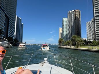 Sunny boat ride down a downtown canal framed by high-rise condos and palm trees, with other yachts ahead and a smiling passenger wearing orange sunglasses on the bow.
