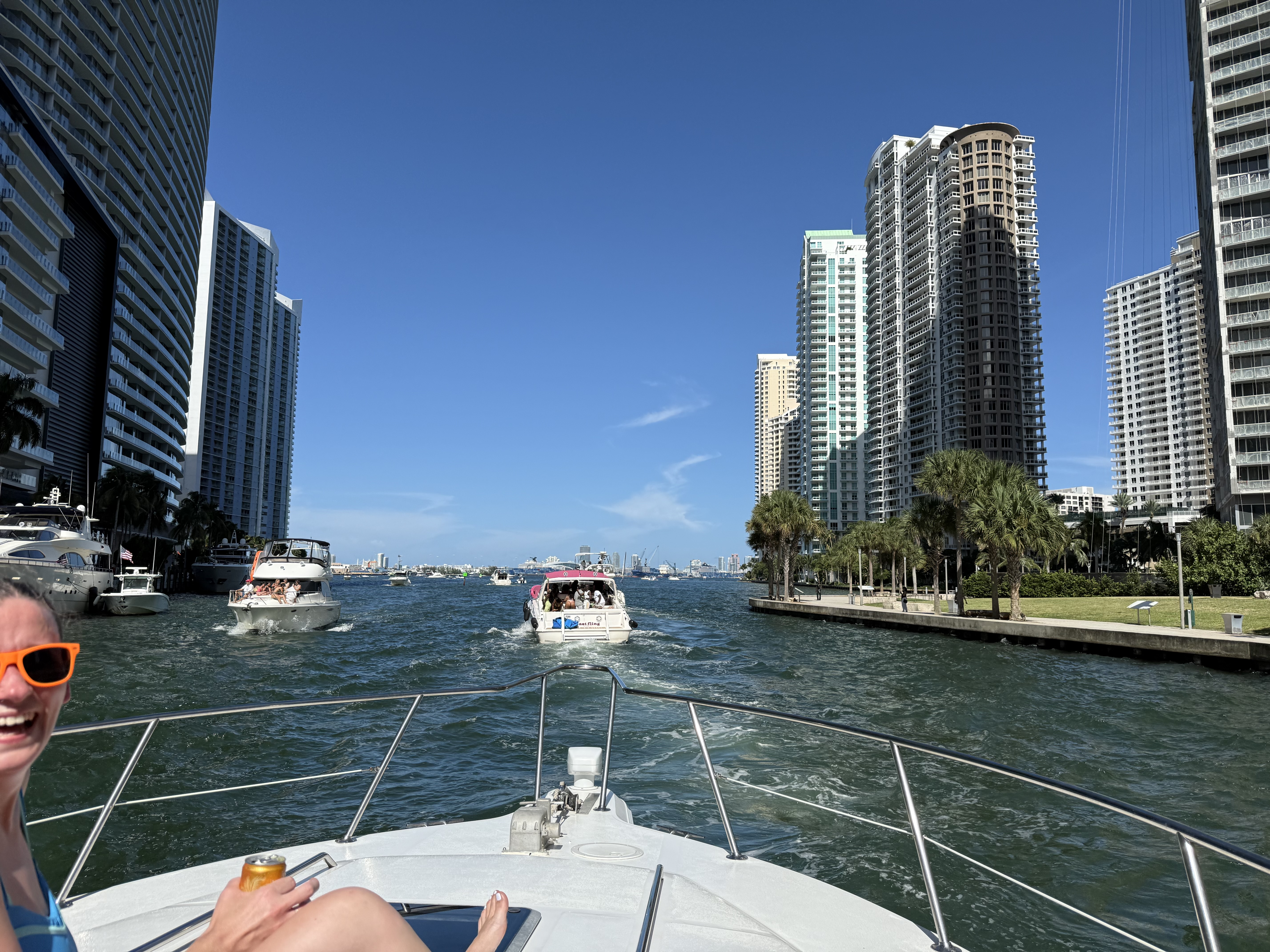 Sunny boat ride down a downtown canal framed by high-rise condos and palm trees, with other yachts ahead and a smiling passenger wearing orange sunglasses on the bow.