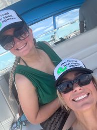 Two smiling women on a boat under blue skies, wearing matching "she found her main squeeze" trucker hats and sunglasses, enjoying a sunny day on the water.