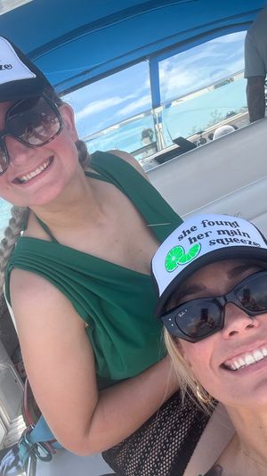 Two smiling women in sunglasses wearing matching trucker hats that say 'she found her main squeeze' with a lime graphic, taking a sunny selfie aboard a boat under a blue canopy.