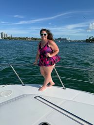 Woman in a purple tropical swimsuit and teal sunglasses posing on the bow of a boat with the Miami skyline, palm-lined shoreline and cruise terminal visible across turquoise water under a bright blue sky.