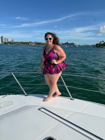 Woman in a purple tropical swimsuit and teal sunglasses posing on the bow of a boat with the Miami skyline, palm-lined shoreline and cruise terminal visible across turquoise water under a bright blue sky.