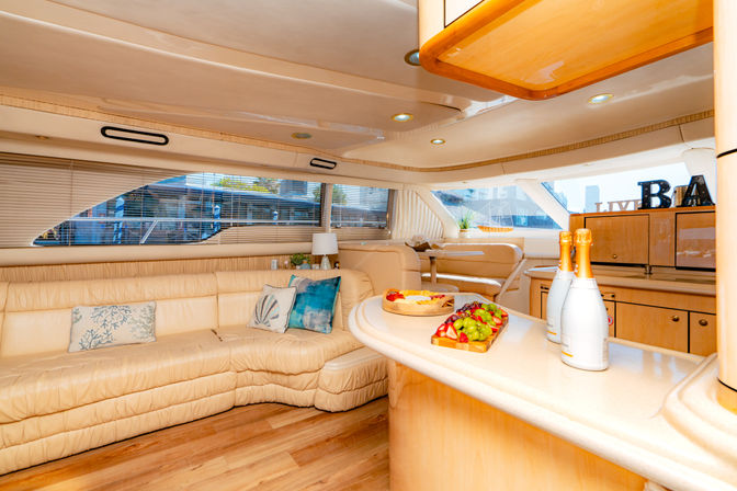 Sunlit luxury yacht salon with cream leather L-shaped sofa and coastal throw pillows, wood floors, curved white countertop topped with champagne bottles and a fresh fruit platter, built-in wood cabinetry and large windows with a marina view.