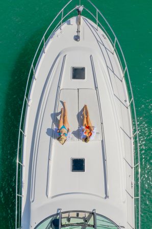 Aerial view of two sunbathers, one in a blue bikini with a straw hat and one in an orange bikini, lounging on the cushioned bow of a white yacht over turquoise sea
