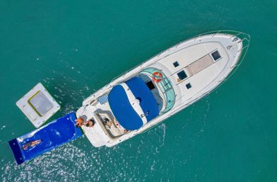 Aerial view of a white motor yacht with a blue sun canopy anchored in turquoise water, passengers on the stern and a bright blue floating mat and swim platform with someone sunbathing.