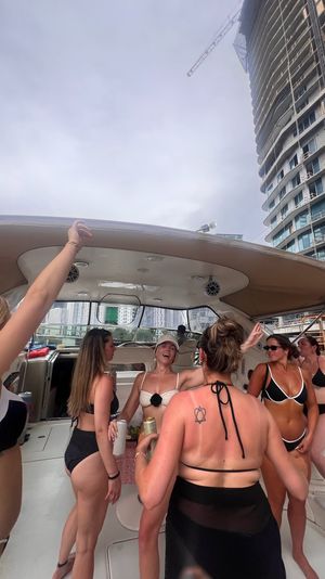 Boat party: group of women in swimsuits laughing and drinking aboard a yacht at an urban waterfront marina with high-rise construction and a crane under an overcast sky.
