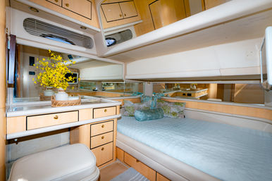 Cozy luxury yacht cabin with light wood cabinets and mirrored walls, pale-blue quilted double berth, floral accent pillows, and a built-in vanity with wicker tray and yellow flowers.