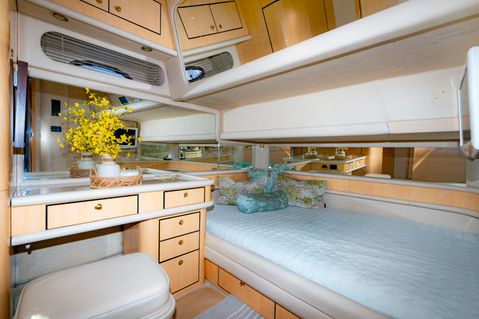 Cozy luxury yacht cabin with light wood cabinets and mirrored walls, pale-blue quilted double berth, floral accent pillows, and a built-in vanity with wicker tray and yellow flowers.