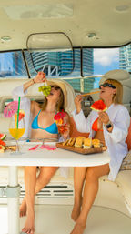 Two friends in bikinis enjoying a sunny yacht brunch—laughing as one feeds grapes and the other bites watermelon—tropical cocktails and snacks on the table with a modern waterfront skyline visible through the windows.