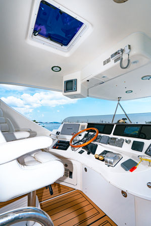 Yacht helm with wooden steering wheel, white leather captain’s chair, multi-screen navigation console and teak-style deck overlooking blue sky and coastal marina