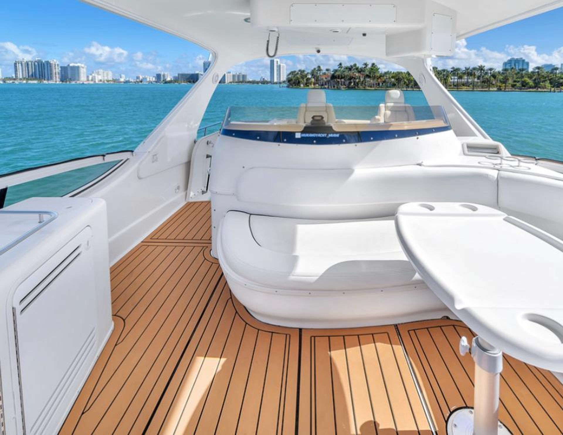Sunny Miami view from a luxury yacht cockpit with teak deck, white curved seating and table overlooking turquoise bay and palm-lined city skyline.