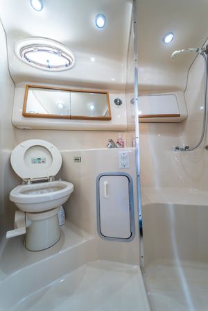 Compact cream-colored yacht bathroom (marine head) with a marine toilet, built-in shower with handheld wand, oval porthole, recessed ceiling lights and a mirrored cabinet.