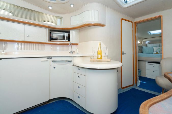 Bright, modern yacht galley and cabin interior with curved white countertop, built-in microwave, sink, champagne bottle and two flutes on a tray, wood trim, skylight and cozy blue-carpeted sleeping berth.