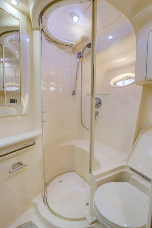 Compact cream-colored marine bathroom on a yacht with curved glass shower enclosure, chrome handheld showerhead, small porthole window and molded toilet.