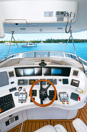Luxury motor yacht helm with wooden steering wheel, dual throttles and navigation screens, overlooking a turquoise coastal marina with anchored boats, tree-lined shore and distant city skyline.