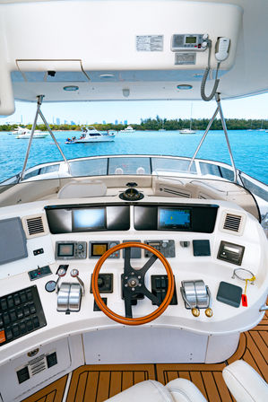 Luxury motor yacht helm with wooden steering wheel, dual throttles and navigation screens, overlooking a turquoise coastal marina with anchored boats, tree-lined shore and distant city skyline.