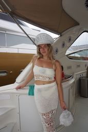 Smiling bride in a sequined “BRIDE” captain hat and white crochet crop top and skirt, holding a fluffy purse aboard a yacht at the marina — playful bachelorette party vibe.