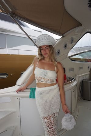 Smiling bride in a sequined “BRIDE” captain hat and white crochet crop top and skirt, holding a fluffy purse aboard a yacht at the marina — playful bachelorette party vibe.