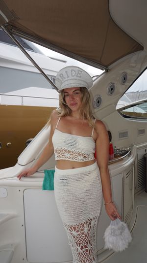 Woman in a white crocheted crop top and skirt wearing a jeweled 'BRIDE' hat and holding a fluffy bridal purse aboard a yacht, with a neighboring boat and marina visible in the background.