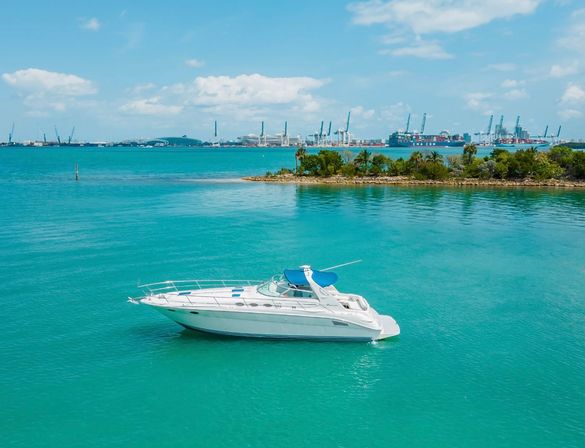 White motor yacht floating on turquoise coastal water near a palm-lined islet with container port cranes on the horizon under a sunny sky