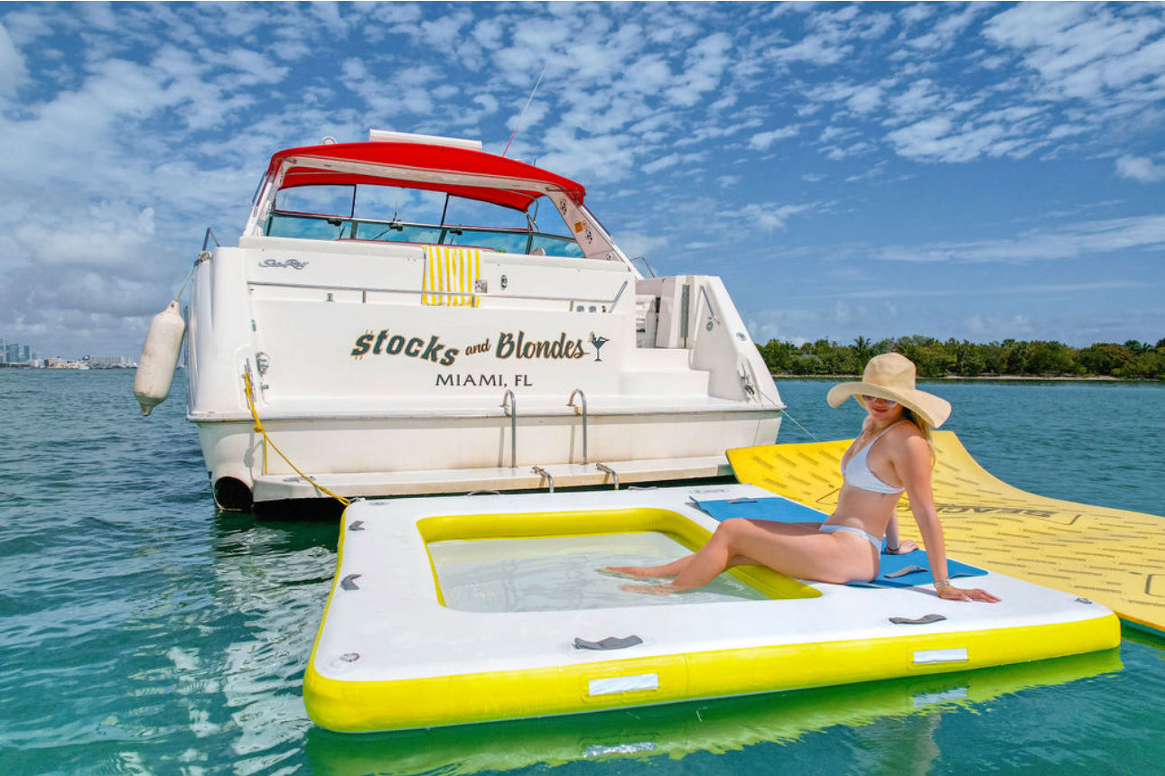 Person in a white bikini and wide-brim sun hat relaxing on a yellow-and-white inflatable platform beside a moored motor yacht in turquoise Miami waters under a bright blue sky