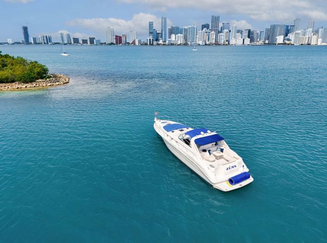White motor yacht with blue canopy on turquoise water near a small mangrove island, Miami skyline in the background under a partly cloudy sky