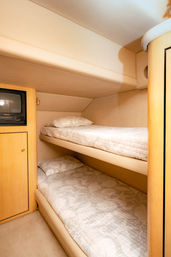Cozy yacht cabin with light wood paneling, stacked upper and lower bunk beds with neutral quilted bedding and a small built-in TV cabinet