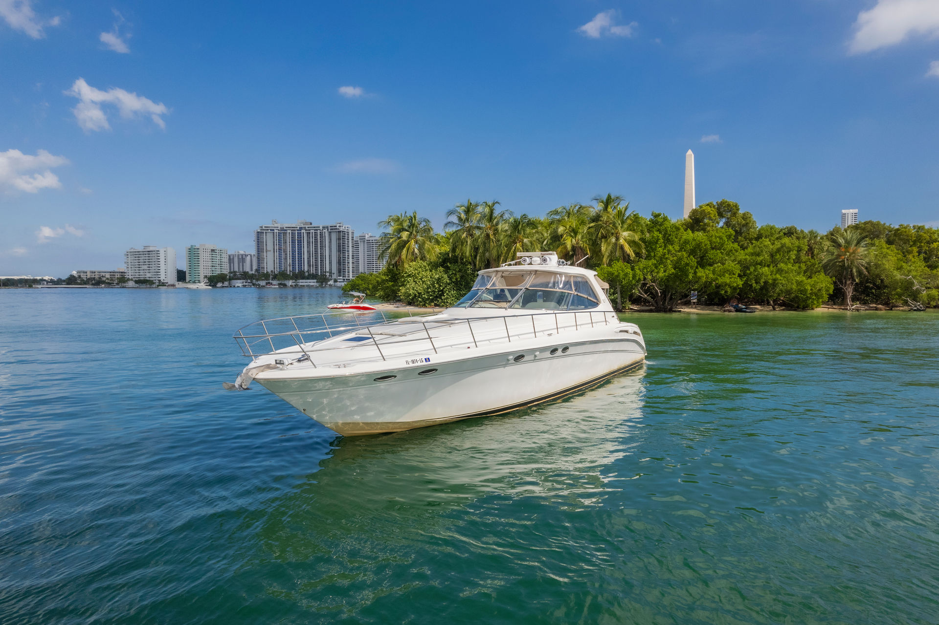 White luxury motor yacht anchored in clear turquoise bay beside a palm‑lined island and high‑rise waterfront buildings under a bright blue sky