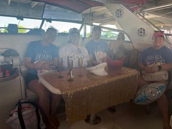 Group of friends celebrating on a yacht docked at a covered marina — gold sequin tablecloth, champagne in an ice bucket, metallic flutes, festive shirts and a balloon for a bachelorette-style party.