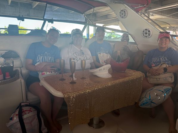 Group of friends celebrating on a yacht docked at a covered marina — gold sequin tablecloth, champagne in an ice bucket, metallic flutes, festive shirts and a balloon for a bachelorette-style party.