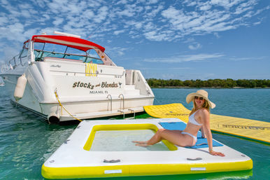 Woman in white bikini and wide-brim hat lounging on a yellow-and-white inflatable platform beside a white yacht in turquoise waters off Miami, Florida — sunny boating scene.