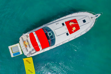Aerial shot of a white luxury yacht in turquoise water with a bright red canopy and sun pad where a sunbather reclines under a straw hat, a yellow inflatable swim mat alongside and cushioned rear deck.