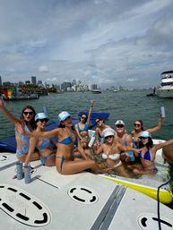 Group of women in colorful swimwear partying on a boat near the Miami skyline, raising drinks and smiling with yachts and coastal cityscape in the background.