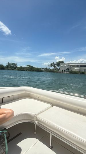 Person in a mint bikini relaxing on a motorboat, holding a drink, cruising turquoise coastal waters past a waterfront building and bridge under a sunny blue sky