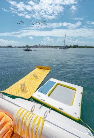 Yellow inflatable water mat and square floating platform tied to a yacht, striped beach towel on the stern, calm turquoise bay with anchored boats, sailboat and birds under a sunny sky.
