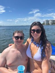 Two people in swimwear and sunglasses on a boat in a sunny bay, blue lei and cup visible, long bridge and waterfront high-rise condos in the background